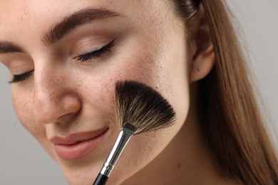 Photo of Beautiful woman with freckles applying makeup with brush on grey background, closeup