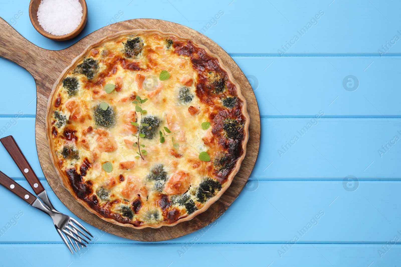 Photo of Delicious homemade salmon quiche with broccoli, forks and salt on light blue wooden table, flat lay. Space for text