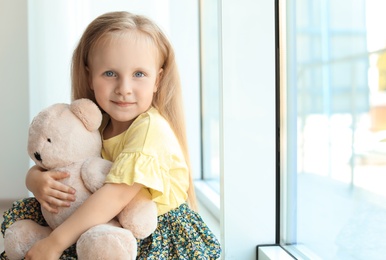 Photo of Pretty little girl with teddy bear near window in room