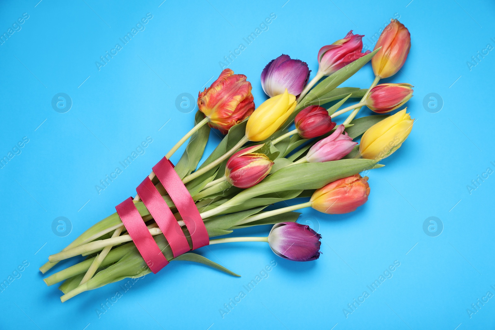 Photo of Bouquet of beautiful colorful tulip flowers tied with red ribbon on light blue background, top view