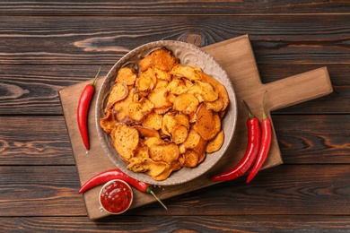 Photo of Board with plate of sweet potato chips, sauce and hot pepper on wooden table, top view