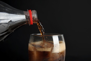 Pouring refreshing cola from bottle into glass with ice cubes on black background, closeup