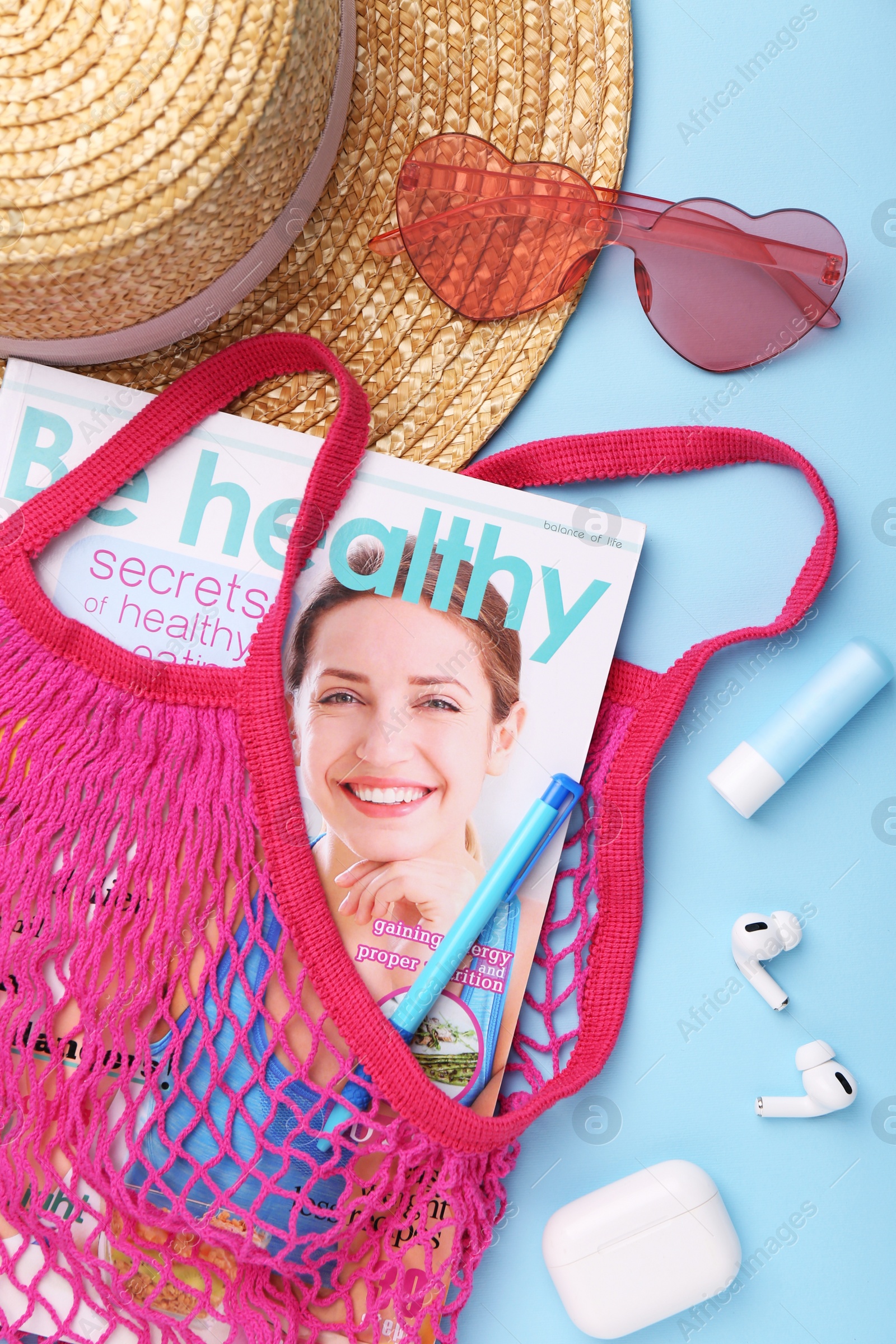 Photo of String bag with magazine, earphones and summer accessories on light blue background, flat lay