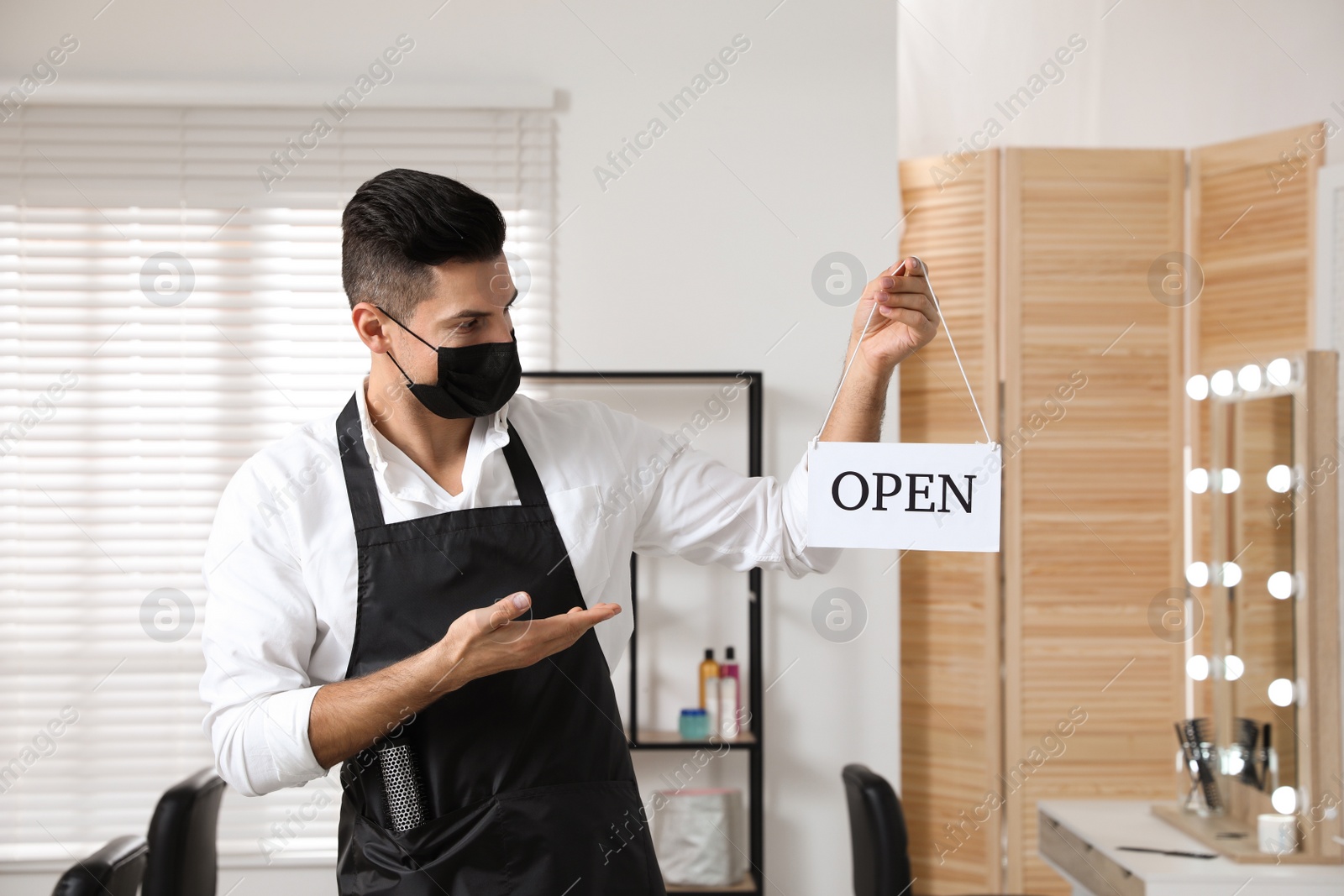 Photo of Professional stylist with protective mask holding Open sign in salon. Hairdressing services during Coronavirus quarantine