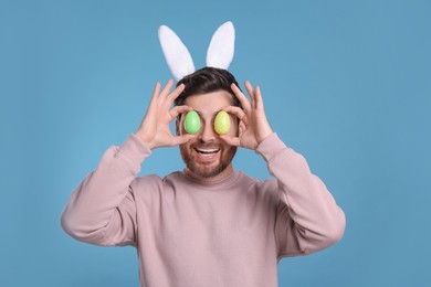 Happy man in cute bunny ears headband covering eyes with Easter eggs on light blue background