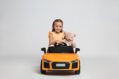 Cute little girl with toy bear driving children's car on grey background