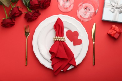 Photo of Place setting with paper hearts, gift boxes and bouquet of roses on red table, flat lay. Romantic dinner