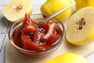 Photo of Tasty homemade quince jam in bowl, spoon and fruits on tiled table, closeup