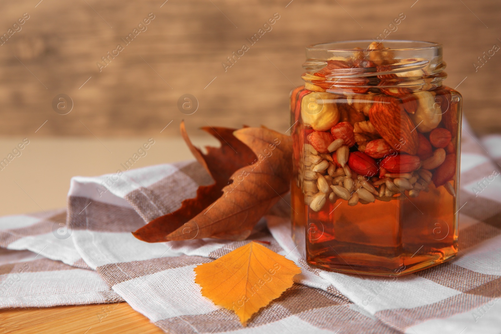 Photo of Different nuts with honey in jar and dry leaves on wooden table. Space for text