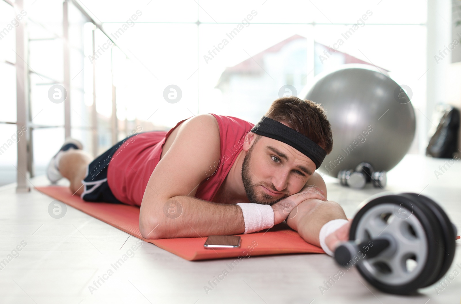 Photo of Lazy young man with abs roller lying on yoga mat indoors