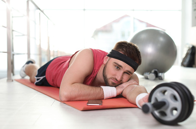 Lazy young man with abs roller lying on yoga mat indoors