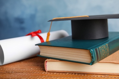 Graduation hat, books and student's diploma on wooden table against light blue background, closeup