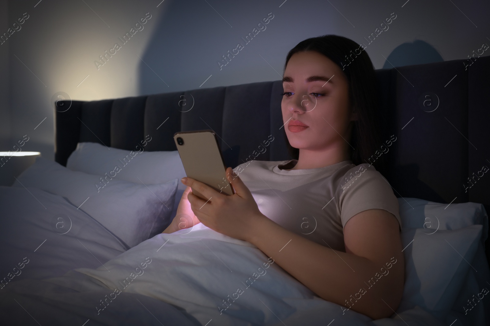 Photo of Young woman using modern smartphone in bed at night. Internet addiction