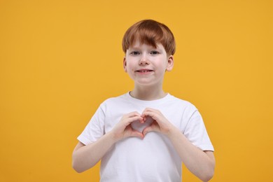 Photo of Cute little boy showing heart gesture with hands on orange background