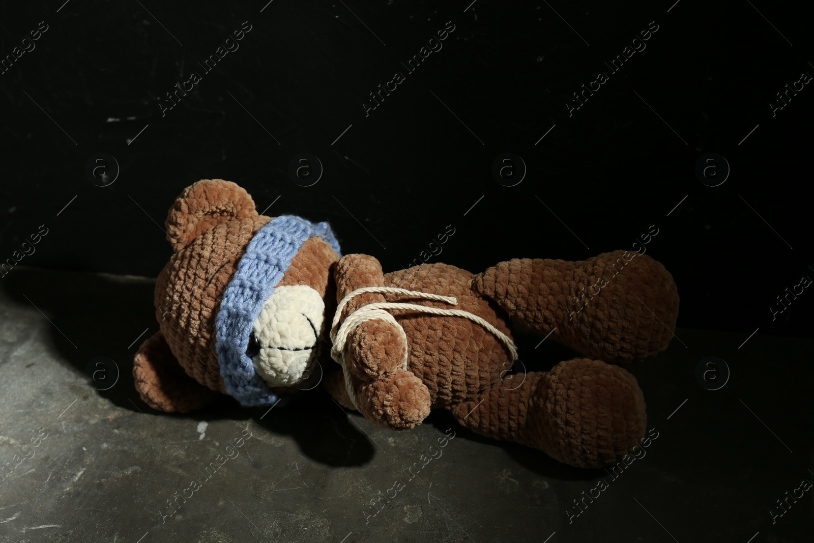 Photo of Stop child abuse. Tied toy bear with taped eyes lying on grey floor against black background