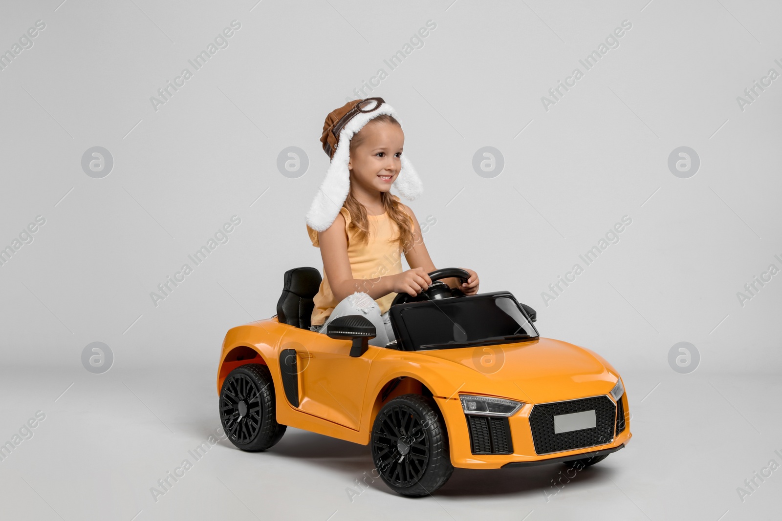 Photo of Cute little girl in pilot hat driving children's electric toy car on grey background