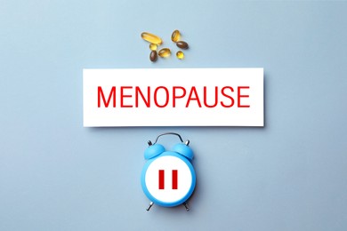 Menopause word, alarm clock with pause symbol and pills on light blue background, top view