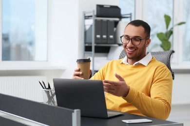 Young man with cup of drink having video chat via laptop in office