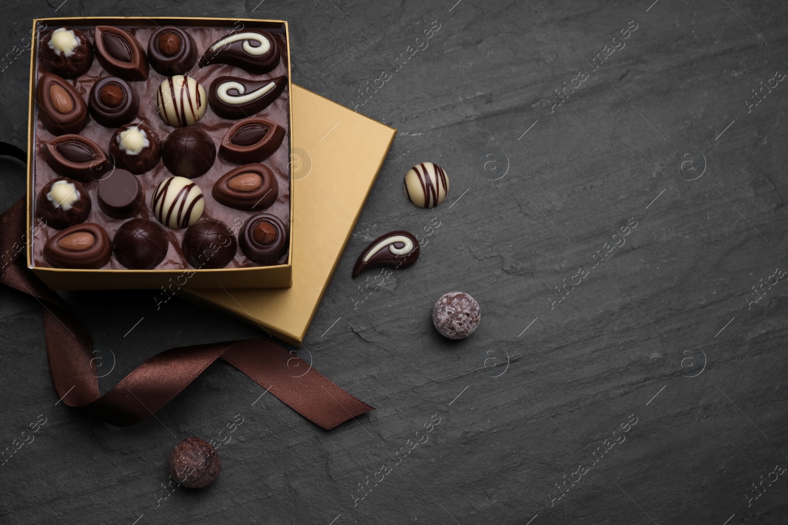 Photo of Open box of delicious chocolate candies and brown ribbon on black table, flat lay. Space for text