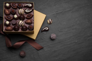 Open box of delicious chocolate candies and brown ribbon on black table, flat lay. Space for text