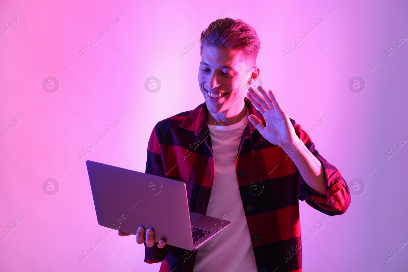 Photo of Young man with laptop talking via video chat on pink background in neon lights