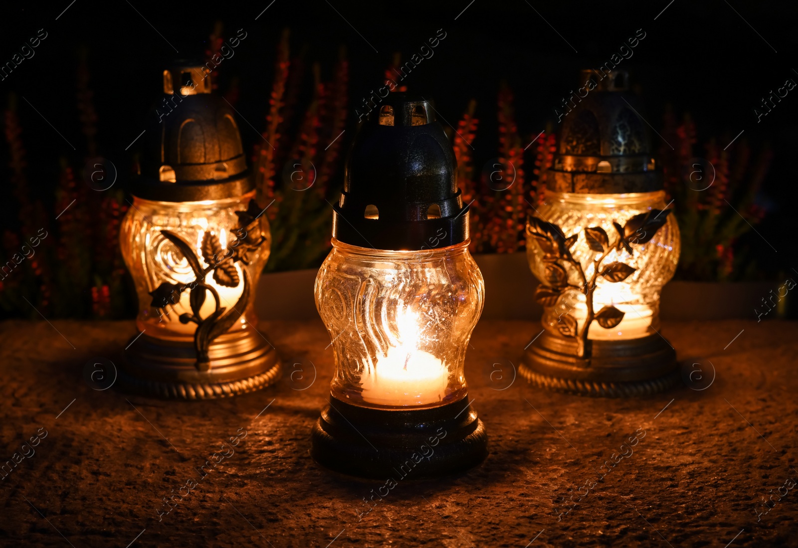 Photo of Different grave lanterns with burning candles on stone surface at night