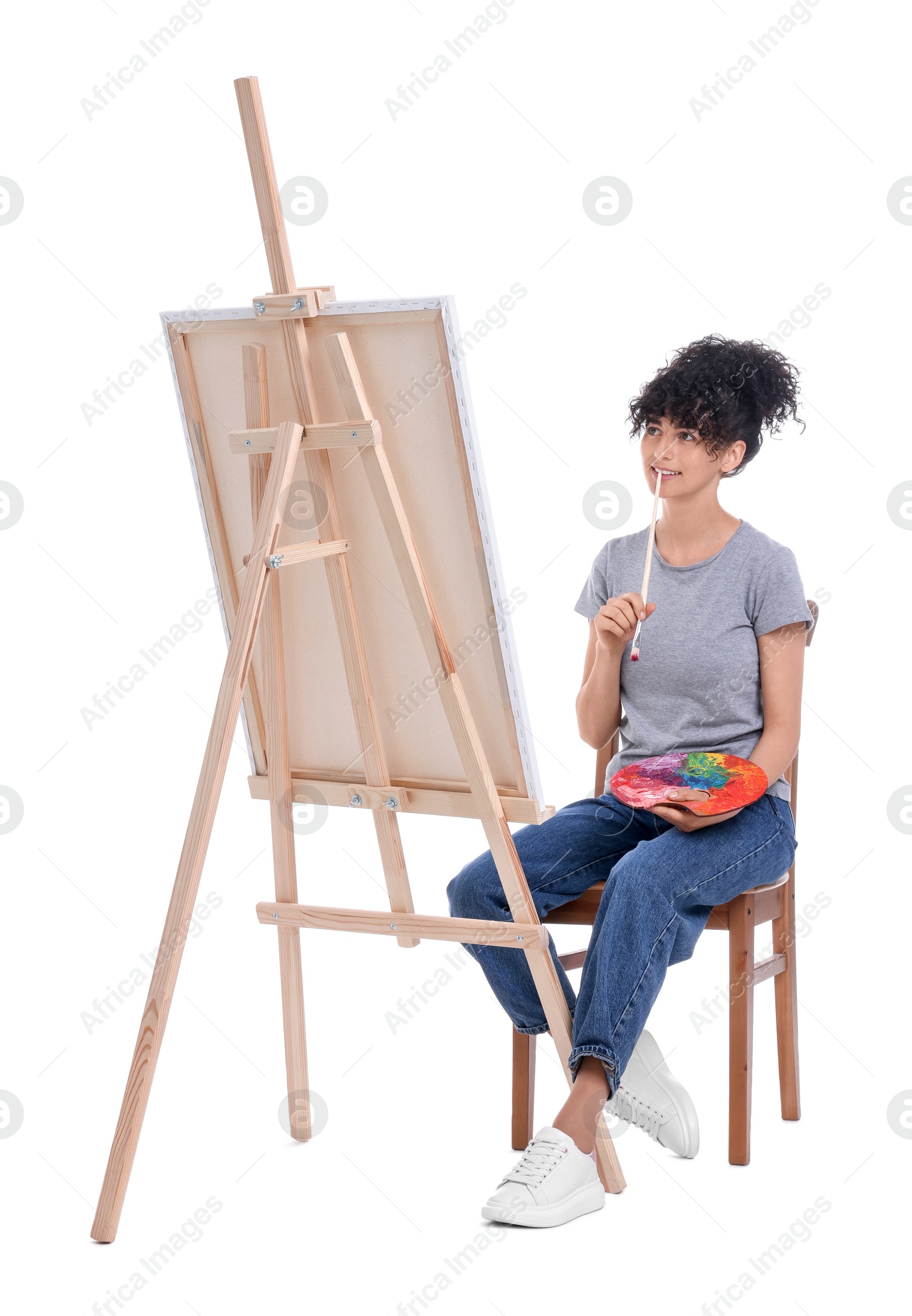 Photo of Young woman holding brush and artist`s palette near easel with canvas against white background