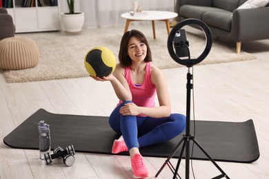 Happy sports blogger holding medicine ball while streaming online fitness lesson with smartphone at home