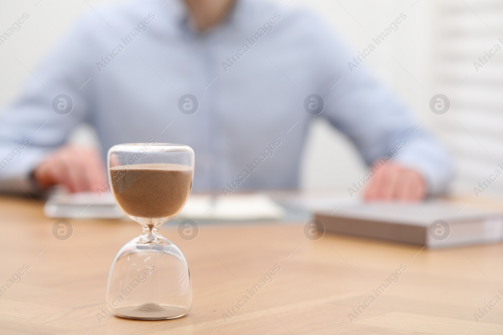 Photo of Hourglass with flowing sand on desk. Man using calculator indoors, selective focus