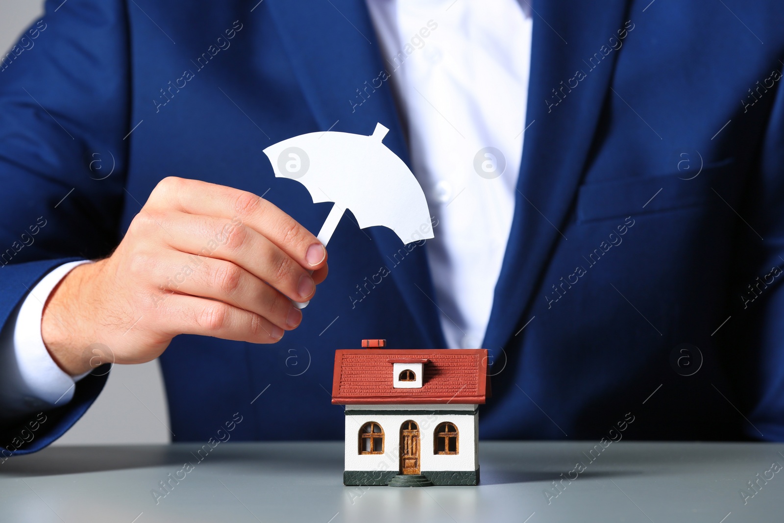 Photo of Man covering house model with umbrella cutout at table, closeup. Home insurance