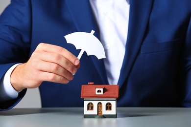 Photo of Man covering house model with umbrella cutout at table, closeup. Home insurance