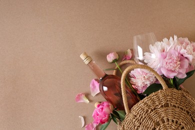 Photo of Wicker bag with bottle of rose wine and beautiful pink peonies on brown background, top view. Space for text