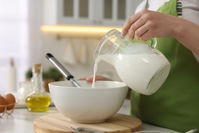 Making bread. Woman pouring milk into bowl at white table in kitchen, closeup