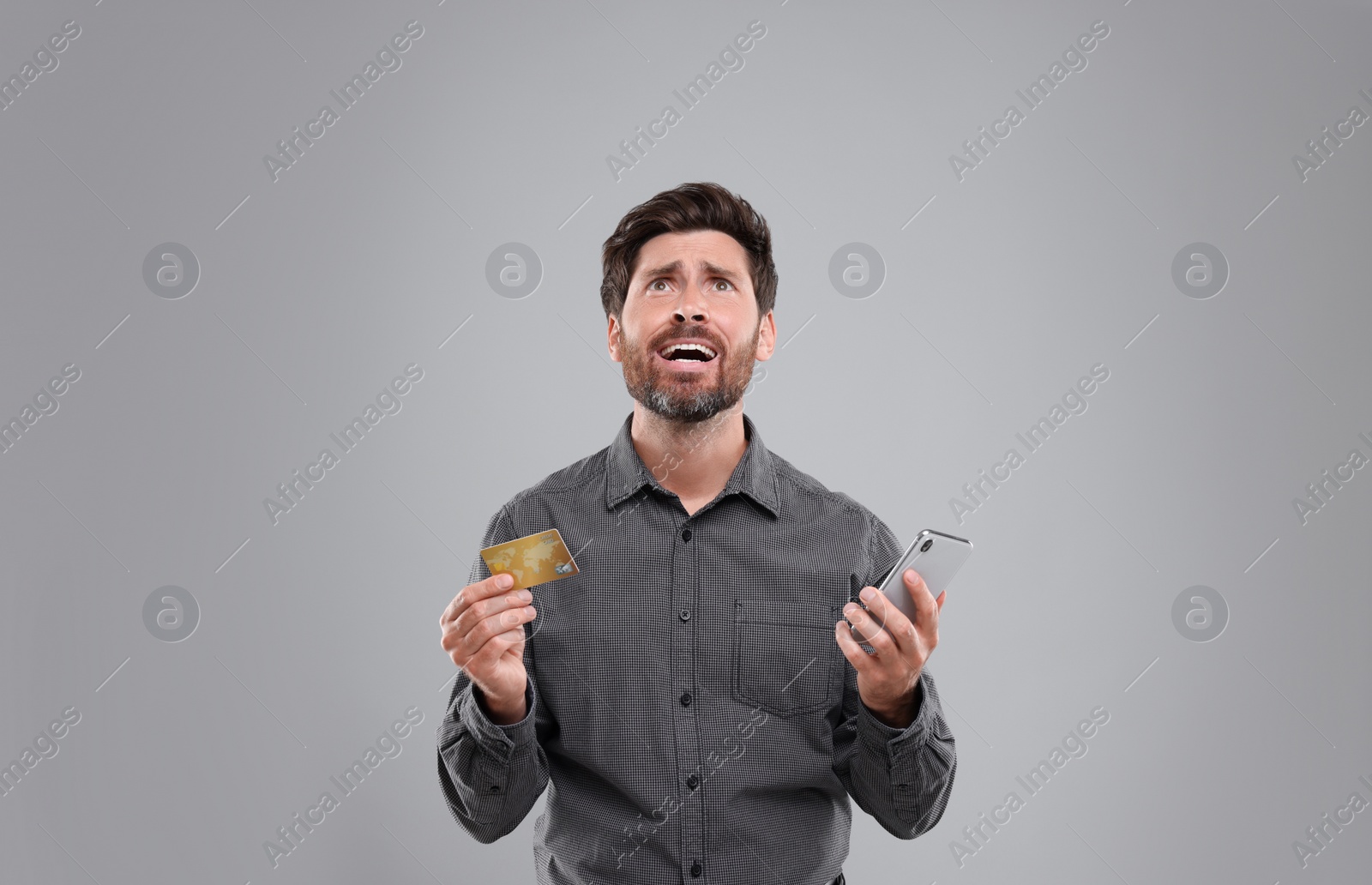 Photo of Emotional man holding smartphone and credit card on light grey background. Be careful - fraud