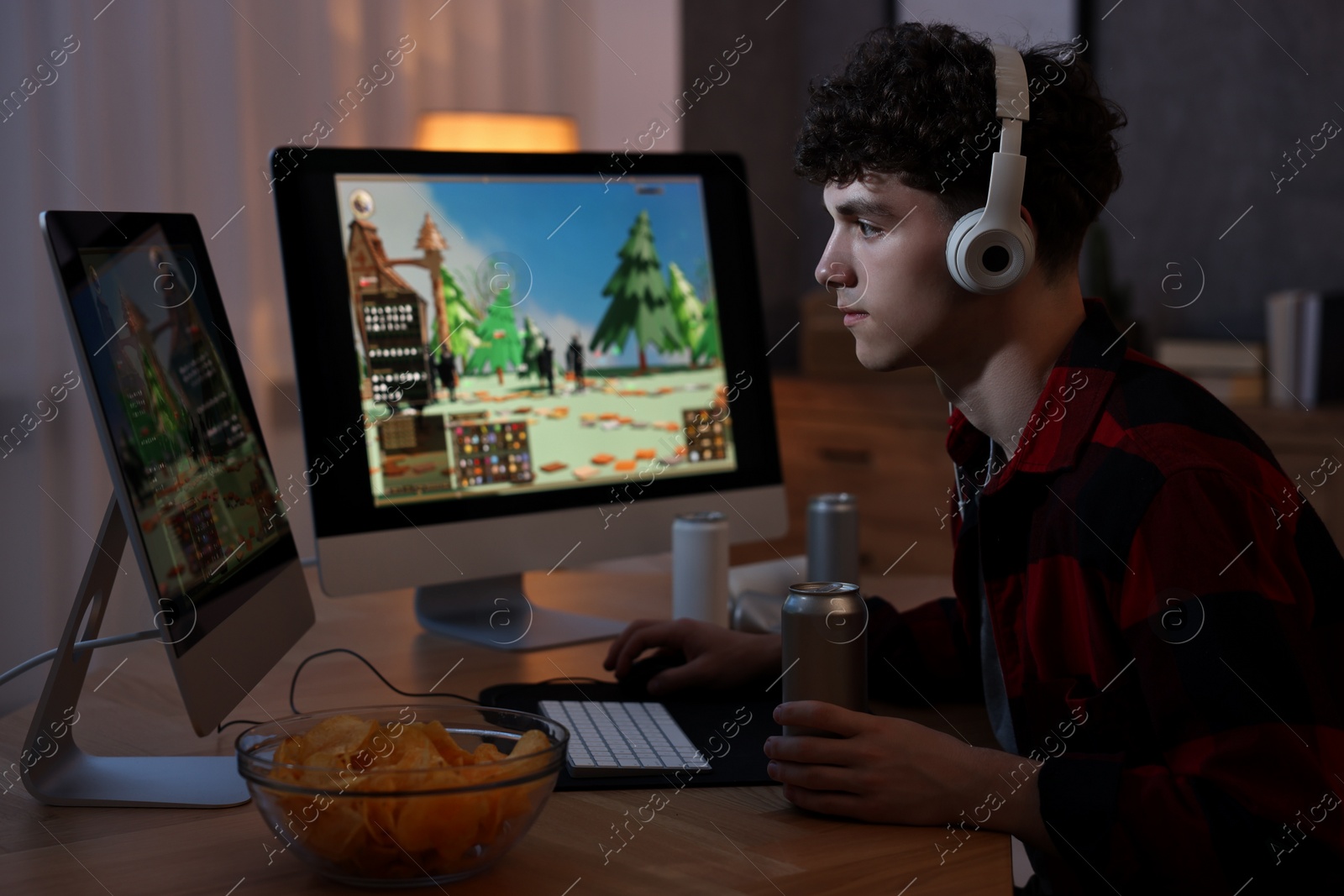 Photo of Young man with energy drink and headphones playing video game at wooden desk indoors