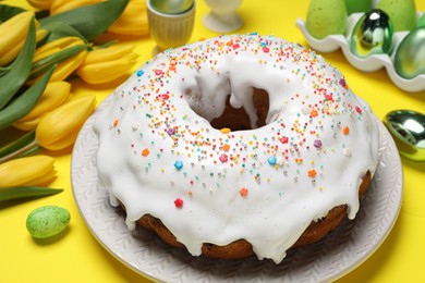 Photo of Easter cake with sprinkles, painted eggs and tulips on yellow background