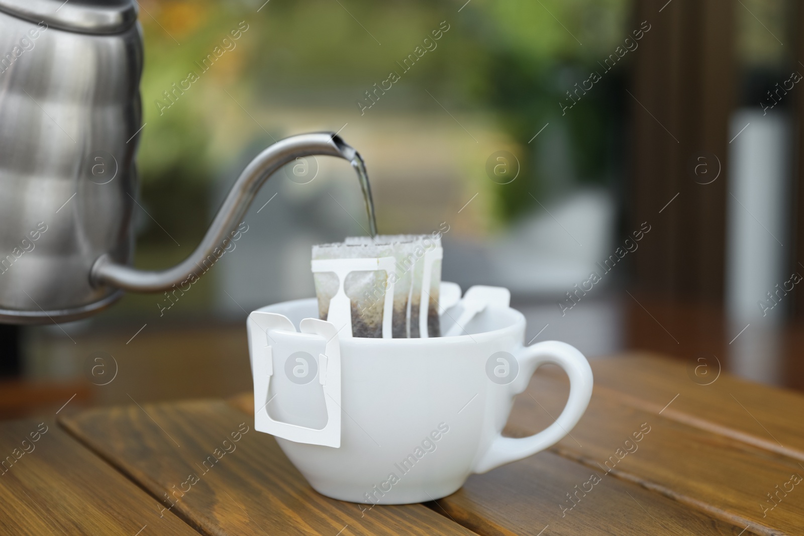 Photo of Pouring hot water into cup with drip coffee bag from kettle on wooden table, closeup