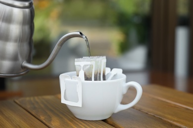 Photo of Pouring hot water into cup with drip coffee bag from kettle on wooden table, closeup