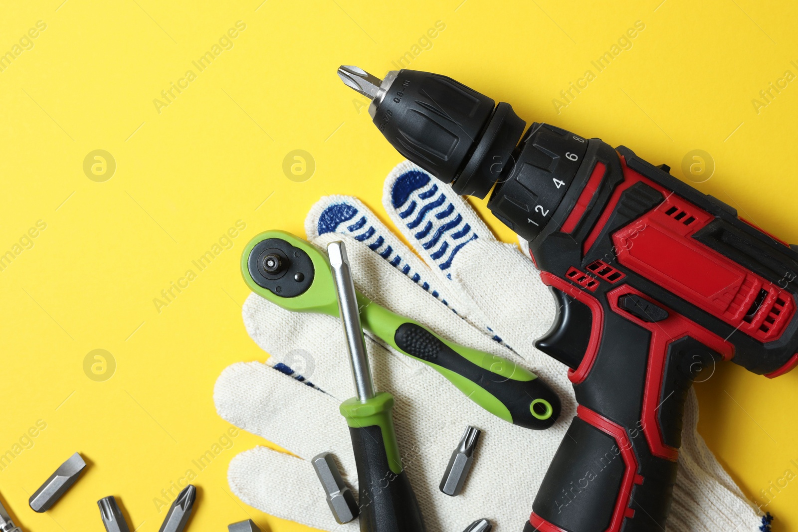 Photo of Electric screwdriver, bit set and gloves on yellow background, flat lay