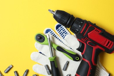 Photo of Electric screwdriver, bit set and gloves on yellow background, flat lay