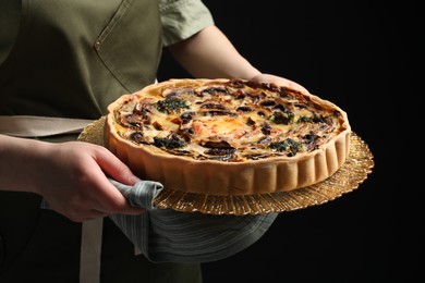 Woman holding delicious homemade quiche with mushrooms against black background, closeup