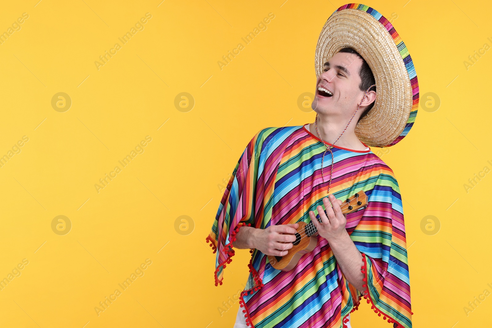 Photo of Young man in Mexican sombrero hat and poncho playing ukulele on yellow background. Space for text