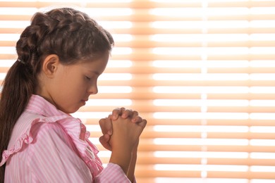 Photo of Cute little girl with hands clasped together praying near window. Space for text