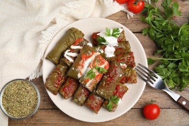 Photo of Delicious stuffed grape leaves with sour cream and tomato sauce on wooden table, flat lay