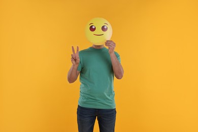 Man covering face with smiling emoticon and showing peace sign on yellow background