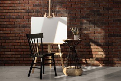 Wooden easel with blank canvas and different art supplies near brick wall in room