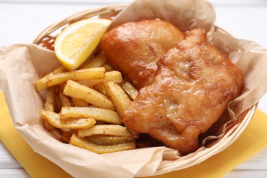 Photo of Tasty fish, chips and lemon in wicker bowl on white wooden table, closeup