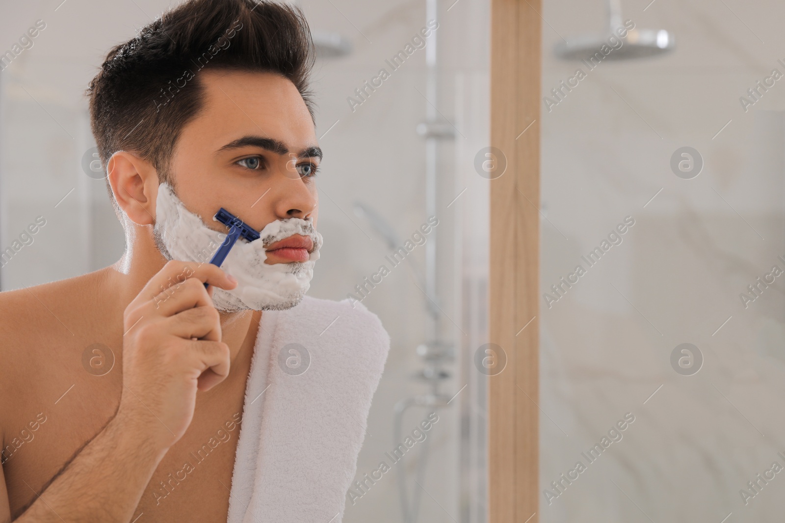 Photo of Handsome young man shaving with razor near mirror in bathroom