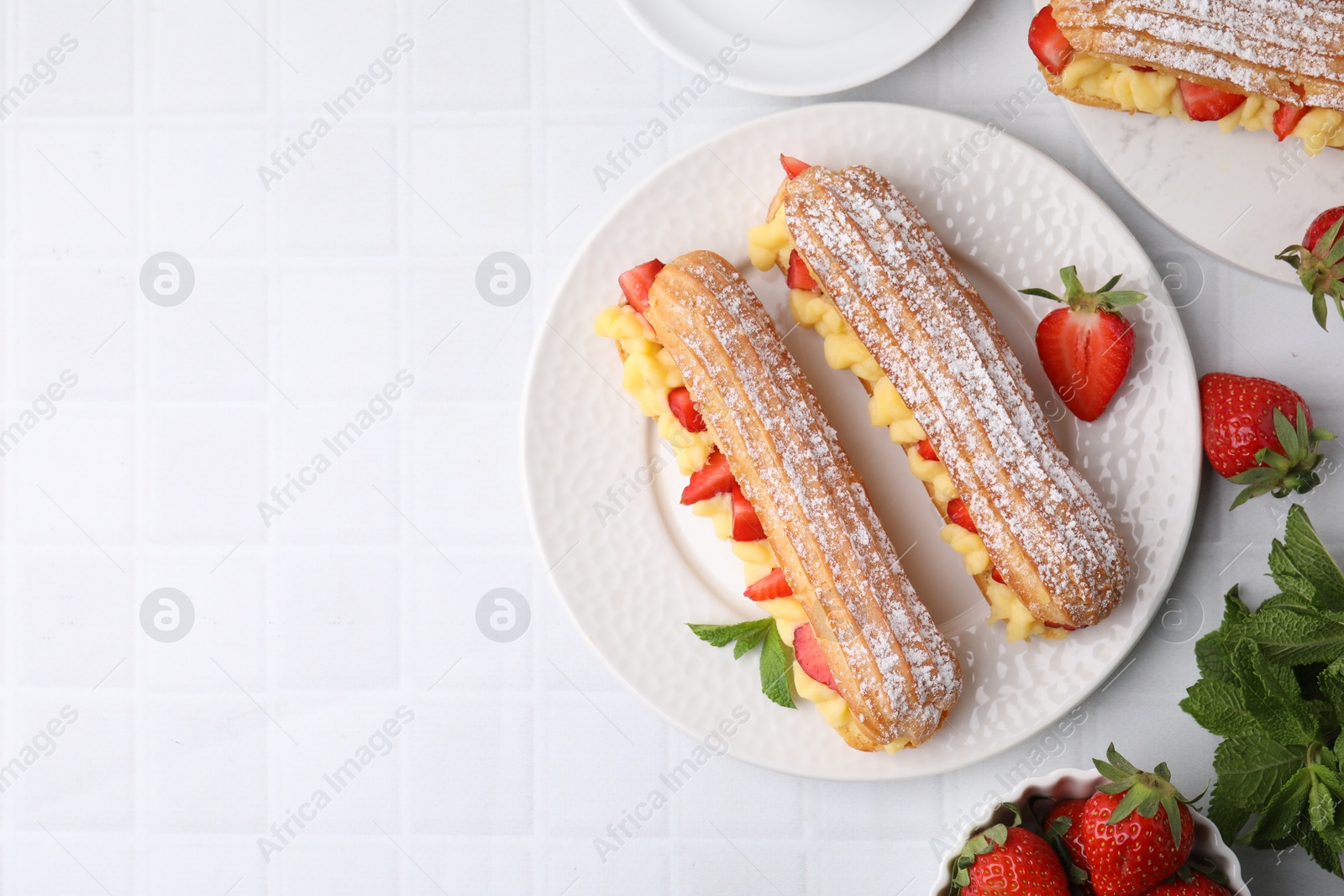 Photo of Delicious eclairs filled with cream, strawberries and mint on white tiled table, top view. Space for text