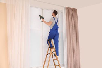 Worker in uniform installing roller window blind on stepladder indoors